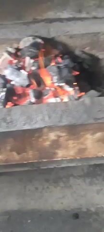 A close-up shot shows burning charcoal in an outdoor grill. The charcoal pieces glow red and orange, with some emitting small flames and smoke. The grill itself appears to be made of metal and concrete. The lighting is dim, suggesting it may be evening or nighttime. There are no people or other objects visible in the frame.