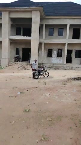 The video shows a man standing next to a parked motorcycle in a dirt area in front of a building under construction. The building is two stories with a dark roof and appears to be a multi-unit structure with rectangular windows. There is another motorcycle parked in front of the building, further to the left. The man is wearing a light-colored shirt and dark pants. The ground is mostly bare dirt with some scattered patches of green vegetation. There is some trash on the ground.  The sky is overcast.