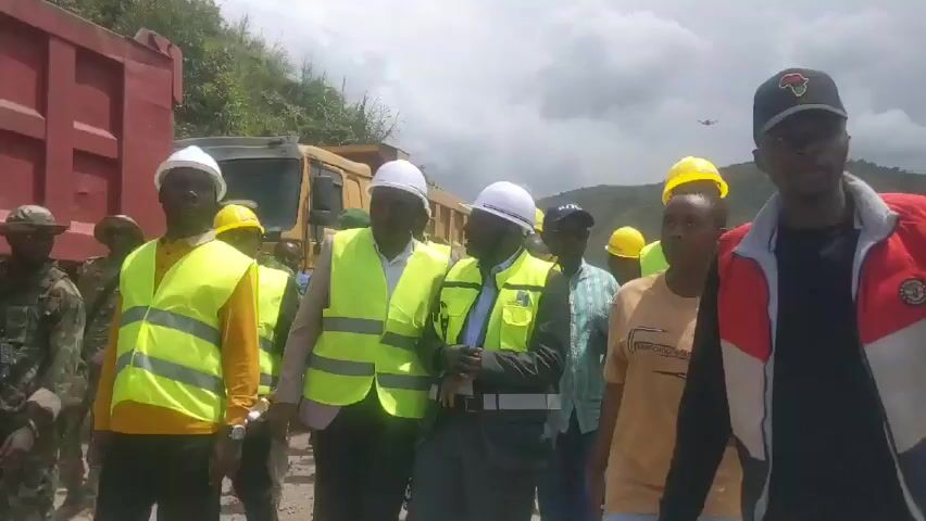 The video depicts a group of approximately twelve individuals standing outdoors. Several people are wearing construction helmets and high-visibility safety vests. Among them are individuals in business attire, including at least one wearing a suit jacket and another with a blue dress shirt and a tie. One woman is wearing a brown t-shirt. Some individuals in the group are wearing casual clothing. The group stands near two large dump trucks, one red and one yellow. In the background, a mountain range is visible beneath a cloudy sky. A small aircraft is visible in the distance. There is a military presence on the left of the group.