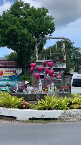 The video shows a roundabout with a fountain decorated with pink heart-shaped balloons. Several jet streams of water are visible from the fountain, and green plants surround the structure. Behind the fountain, a large tree with green leaves stands tall. In the background, a white van is parked, and a person wearing a white helmet is riding a motorcycle past the roundabout. Another motorcycle and a yellow car are also visible in the distance. The sky is cloudy.