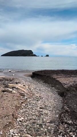 The video shows a view from a beach looking out towards a body of water. A small stream of clear water flows through a pebble-filled channel toward the water. The beach consists of sand, pebbles, and darker patches of soil. In the distance, an island is visible, dark in color and topped with greenery. The sky is light blue with fluffy white clouds. The water is calm and still. There are no people visible in the video.