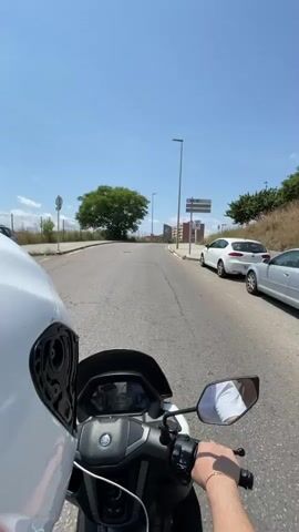 The first-person perspective video shows a person riding a white Yamaha scooter on an empty road on a bright, sunny day. The rider wears a white t-shirt and a silver bracelet. In the background, there are buildings, trees, and street signs. The road curves gently uphill. A white car is parked on the right side of the road at the beginning of the video.