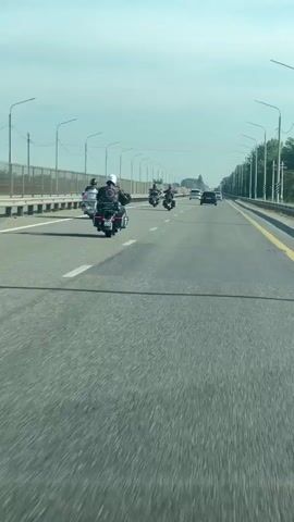 A group of at least seven motorcycles and two cars are traveling down a multi-lane highway under a clear sky. The motorcycles are in a staggered formation, with some in the left lane and others in the right lane. One motorcycle in the foreground is in the right lane and is the closest to the camera. The cars are further down the highway, also in the right lane. The highway is lined with streetlights and a metal fence on the left side. Trees are visible in the distance on the right side of the highway. The video appears to be shot from the perspective of someone driving a vehicle behind the motorcycles.