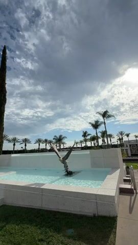 The video pans from a fountain with a bird sculpture to a row of palm trees and modern buildings. The sky is partly cloudy. In the foreground, there is a well-maintained lawn. There are a few people visible walking near the buildings in the distance. The architecture appears contemporary, with glass facades and sharp, angular designs. Two tall, slender trees stand prominently in the mid-ground. The fountain is modern and geometric, with clear blue water. The overall impression is one of a clean, modern, and well-landscaped environment.