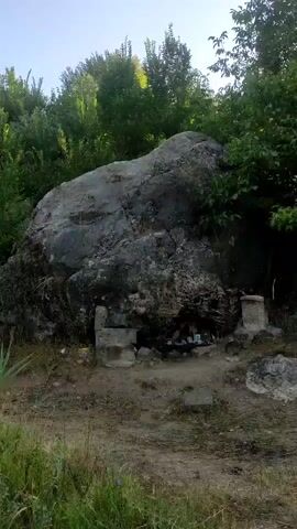 The video shows a large rock formation with a small opening or alcove at its base. Inside the alcove, there are several framed pictures, some bottles, and what appears to be offerings of some kind. There are also some stone structures around the alcove, resembling small altars or benches. The area is covered in dirt and dry vegetation, with trees and foliage in the background. The lighting suggests it is daytime. There are no people visible in the video.