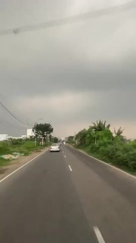 A point-of-view shot from a moving vehicle captures a two-lane asphalt road under a heavily overcast sky. The vehicle travels straight ahead on the right side of the road, marked by a dashed white line. A white car drives ahead in the same lane, and an auto-rickshaw follows it. Another auto-rickshaw approaches in the opposite lane. Green foliage, including trees and bushes, lines both sides of the road. On the left, power lines are visible, with buildings in the background. On the right, a multi-colored fence is visible behind the foliage.