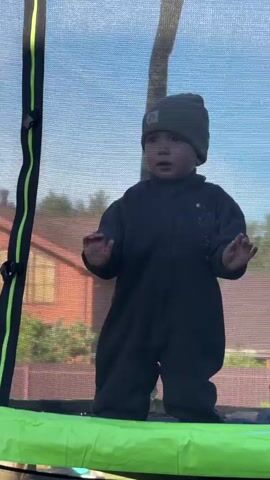 A toddler stands inside a trampoline enclosure wearing a gray beanie and onesie. He has his hands raised and is looking directly at the camera, which is positioned just outside the trampoline's netting. A section of a house is visible in the background, along with foliage and a fence. Another person can be seen slightly to the right of the toddler, also inside the trampoline. The sky is light blue, indicating daytime.