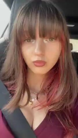 A young woman is in the passenger seat of a car, wearing a seatbelt and recording a video. She has medium-length brown hair with bangs and pink highlights framing her face. She is wearing a maroon V-neck shirt and multiple rings and necklaces. She looks directly at the camera and smiles. The interior of the car is visible, with dark leather seats. The angle of the video shifts during the recording.