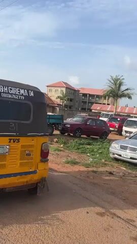 The video shows the view from a moving motorcycle, passing by people and buildings in an outdoor environment during daylight. The motorcycle passes a yellow three-wheeled vehicle, a “keke,” with a black hood. The keke has writing on the back that says "BBH GIREMA 04" and a phone number. The motorcycle then passes a sign that reads "SALLY INT’L ACADEMY ABUJA ROAD NORTH RIGASA KADUNA." A group of women wearing dark clothing and head coverings stand near the sign. The motorcycle continues past the school, where a man in a blue outfit leads a woman in a full black abaya across the sidewalk.