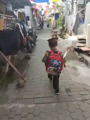 The video shows a young boy walking away from the camera down a narrow, paved alleyway. He is wearing a dark hat, a light-colored shirt, dark pants, and a red backpack with a cartoon design. He is carrying a small Indonesian flag in his right hand and appears to be holding something else in his left hand. The alley is lined with buildings and houses on both sides, with laundry hanging from clotheslines. Several motorcycles are parked along the left side of the alley. Decorative flags are strung across the alley overhead. The ground is paved with interlocking bricks. The overall environment suggests a residential area, possibly in Southeast Asia.