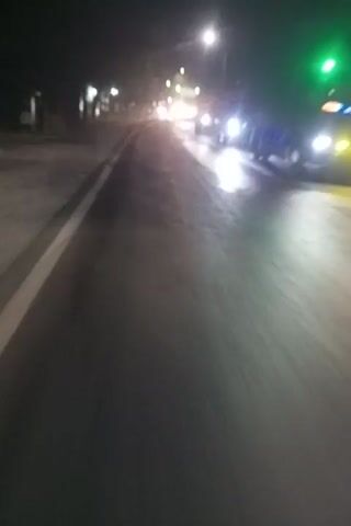 The video shows a nighttime view from a moving vehicle looking forward and slightly to the left. The road surface is visible, wet and reflective, with some streaks of light. On the left side of the road, there are streetlights, some with green or blue illumination, and distant lights that suggest a town or city. There are also trees and the blurred shapes of buildings or structures. The camera is angled such that the scene appears tilted. The vehicle appears to be moving at a moderate speed. There are no visible people.