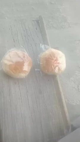 The video shows two round, bread-like objects, each individually wrapped in clear plastic. They are positioned on a weathered wooden surface, possibly a plank or bench. The surface appears to be outdoors or in an unfinished space, as indicated by the rough texture and the muted, dusty lighting. The objects are light brown on top and lighter underneath. The right object has a slight reddish tinge visible through the plastic. The camera angle is overhead, looking down at the objects. The background is a blurred, light gray, suggesting an out-of-focus environment. There are no discernible actions or interactions taking place; the objects are static.
