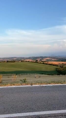 This is a vertical, medium-angle, full-shot video of a rolling countryside landscape under a bright blue sky with scattered clouds. In the foreground, there is a paved road with a white line marking the edge. Beyond the road, a field slopes downwards, dotted with numerous round hay bales. The middle ground features more rolling hills with patches of green and golden-yellow fields, indicating agriculture. A small body of water, possibly a pond or lake, is visible in a dip between the hills. In the distance, a hazy panorama of more hills and distant mountains can be seen, along with scattered buildings. The overall lighting suggests it is late afternoon or early evening, with the sun casting a warm glow on the landscape. No people or animals are visible in the video.