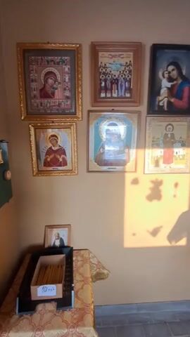 The video shows the inside of what appears to be a small chapel or religious space. The walls are painted a light beige or tan color, and the ceiling has some blue trim. Various religious icons and framed images are hung on the walls. There's a table covered with a decorative cloth, holding more religious images and papers.  Near the first wall is a box containing candles.  A window shows an outside scene with snow on the ground. There is a simple wooden desk with papers on it, and a brown chair. A golden candlestick stands near the window. The overall impression is a quiet, contemplative space. There are no people visible in the video.