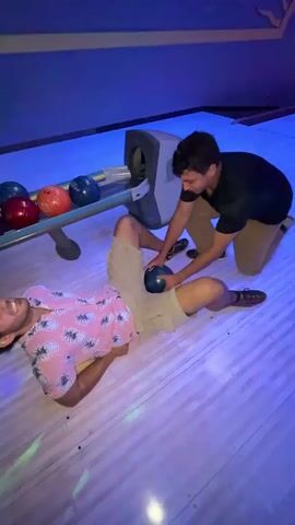 Two young men are in a bowling alley. One man is lying on his back on the bowling lane wearing a pink shirt with a pineapple pattern and tan shorts. The other man is kneeling beside him on the lane, holding a bowling ball and placing it on the first man's stomach. In the background, a third man is standing and holding another bowling ball. The bowling pins are visible in the background. The lighting is dim and colorful, with blue and purple lights reflecting off the lane.