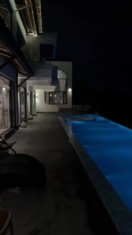 The video depicts a nighttime view of a pool area attached to a building. The pool is illuminated with blue light, and the building has white exterior walls with glass windows. The view pans toward the pool's edge, revealing a dark background possibly indicating an open landscape or body of water. There are a few distant lights visible on the dark horizon. Upbeat music is playing in the background.