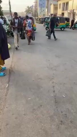 The video depicts a bustling street scene in Alheri, Nigeria, under clear, sunny daylight. Numerous people are seen walking along a paved street and sidewalk. In the foreground on the left, a person wearing a yellow garment and a black overgarment is partially visible from behind. Another individual, dressed in a brown jacket and light-colored trousers, walks away from the camera, carrying a black item over their arm.

Throughout the midground and background, other pedestrians, including men and women of various ages, are moving in different directions. Some individuals are carrying bags or other items. Several three-wheeled vehicles, commonly known as Keke Napep or auto-rickshaws, are visible on the street, suggesting active public transport. At least two of these vehicles are green or blue in color. Faint text, including "PT," can be seen on the side of one of the tricycles. Buildings are visible along the street in the distance, indicating an urban or semi-urban setting. The atmosphere appears to be that of a busy local thoroughfare.