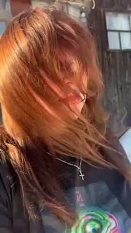 A woman with red hair is outside in front of a building. She wears a black t-shirt and a silver cross necklace. The sun is shining on her face and the wind is blowing her hair. She looks around. The camera is filming at a low angle.