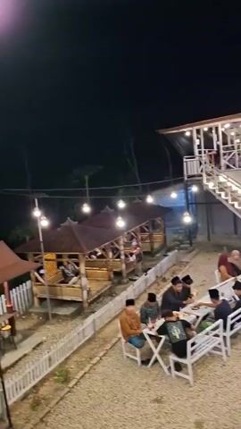 The media file captures an outdoor evening scene at a cafe or restaurant in Lantek Barat, Indonesia. The setting appears to be a gravelly outdoor seating area with numerous white picnic tables and benches. String lights are strung overhead, casting a warm glow, and larger, decorative pendant lights hang above some of the tables. The atmosphere suggests a lively but relaxed environment.

Several groups of people are seated at the tables, engaged in conversation and dining. The patrons are dressed casually, and many appear to be men. Some individuals are wearing traditional Islamic attire, such as peci caps. In the background, a two-story building with a modern design serves as the main establishment, featuring an upper-level balcony and a ground-floor area with more seating and what appears to be a counter or bar. An external staircase connects the two levels.

Adjacent to the seating area, a road is visible with several motorcycles parked along the side. A few people are walking on the road, and one person is riding a motorcycle. The overall impression is of a popular local gathering spot, likely a cafe or eatery, where people are enjoying their evening. The time of day is clearly nighttime, indicated by the darkness of the sky and the illuminated lights. The weather appears to be clear, as there are no visible signs of rain or strong wind. The scene conveys a sense of community and social interaction. No legible text is visible in the video.