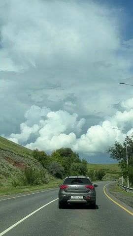 The video shows a car driving on a paved road. The sky is partly cloudy with large, white cumulus clouds. The road is a two-lane highway with a white solid line on the left and a yellow solid line on the right. Green hills with trees and vegetation are visible on the left side of the road. On the right side of the road, there are more trees and lampposts. Another car is seen driving in the opposite direction. The rear of the car in front of the camera is visible, showing a license plate. The weather appears to be partly sunny.