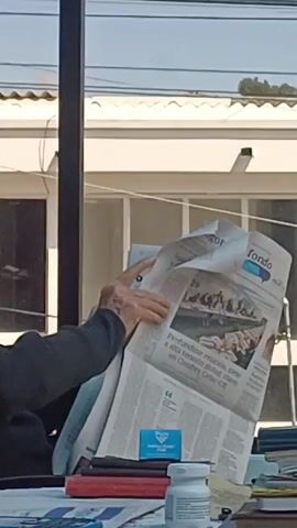 An elderly man is seated at a table outside, reading a newspaper. He wears a black pinstripe hat, a black sweater, and a plaid collared shirt. His hands are visibly wrinkled. On the table is a stack of what appears to be books, a business card, and a bottle of pills. The setting appears to be an outdoor patio, with a white building and power lines visible in the background. The man is focused on reading the newspaper.