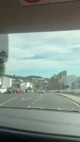 The video shows a view from inside a car driving uphill on a road in a city. Palm trees line the right side of the road. A highway with traffic is visible below the road, and a mountain is visible in the background. The sky is partly cloudy. Several cars are visible on the road. The focus is on the view ahead of the car. The car is moving at a slow pace.