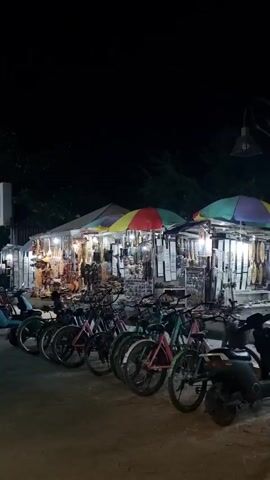 At night, a line of rental bicycles sits parallel to street vendor stalls. The stalls have colorful awnings and are selling various handcrafted souvenirs, clothing, and jewelry. People are riding bicycles and mopeds down the road along the stalls. The night sky is dark and the stalls are illuminated by lanterns and lights.