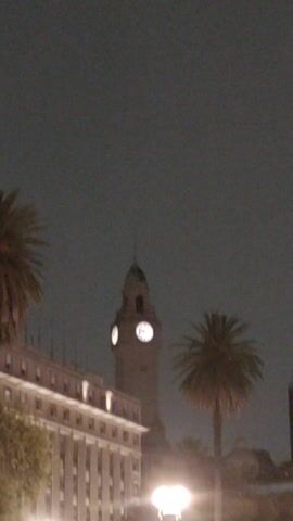 The video pans across a nighttime urban scene, featuring a clock tower with illuminated clock faces, a multi-story building, and palm trees. The sky is dark, suggesting it is late evening or night. Streetlights are visible in the foreground, providing illumination. The buildings and trees are silhouetted against the dark sky. The camera slowly pans to the right.