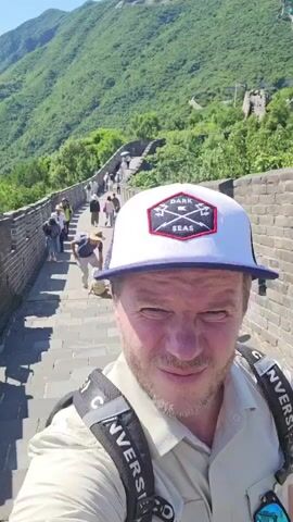 A man wearing a white and blue baseball cap with a black and red logo, a light-colored button-up shirt, and a backpack with black straps is taking a selfie. He is standing on a stone staircase that leads up a section of the Great Wall of China. Behind him, the wall stretches into the distance, lined with tourists walking and sitting. The surrounding landscape is hilly and covered in lush green vegetation. The man is smiling at the camera.
