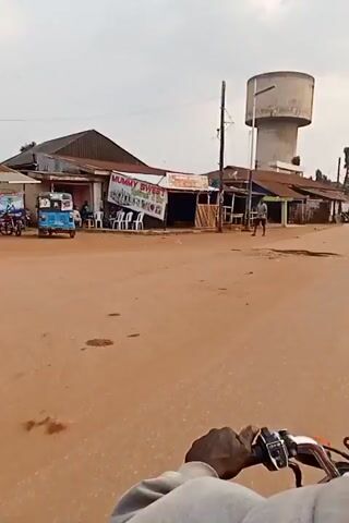 A motorcycle is traveling on a paved road in what appears to be a small town. On either side of the road are various small businesses or shops, some appearing to be made of corrugated iron or other basic materials. A water tower is visible in the background. There are a few people walking or standing near the shops, and another motorcycle is seen in the distance. The weather appears to be overcast, and the road is predominantly reddish-brown in color.