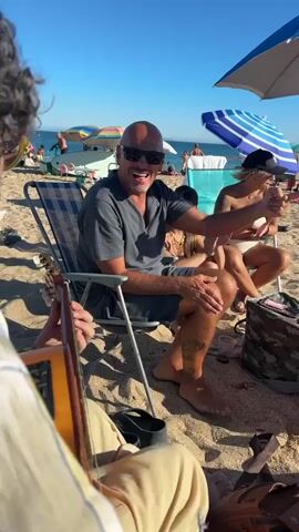 The video shows a group of people on a sunny beach. In the foreground, a person is holding an acoustic guitar, their hand and arm visible. In the mid-ground, a bald man wearing sunglasses and a dark grey short-sleeved shirt sits in a blue and white striped beach chair, smiling and gesturing with his right hand. He has tattoos on his leg. To his right, a woman wearing a white bikini and a black baseball cap sits in another beach chair, also smiling. Behind them, other beachgoers and umbrellas are visible. The scene is set on a sandy beach with the ocean and sky in the background.