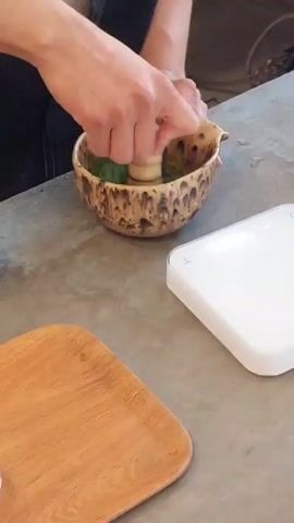 Here is a concise description of the video: 

The video shows a person mixing ingredients in a mottled ceramic bowl with a wooden pestle. To the side is a digital kitchen scale displaying a weight measurement and a small, wooden cutting board. The person is wearing a dark apron and is working on a gray countertop. The action focuses on the mixing process.