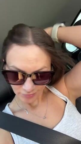 A woman with long brown hair wearing sunglasses and a white tank top is in the passenger seat of a car. She is adjusting her hair and puckering her lips. A seatbelt is visible across her chest. The video is shot from a close-up perspective, showing her face and upper body. The background is blurred, but appears to be the interior of a car with the sky visible through the windows, suggesting she is in motion.