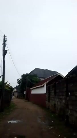 The video shows a narrow, unpaved alleyway lined with buildings and a brick wall. The sky is overcast, suggesting a cloudy day. A wooden utility pole with wires runs along the left side of the alley. In the background, several buildings of varying architectural styles are visible, including one with a dark, angled roof. The alley is damp, with puddles of water visible on the ground. The overall impression is one of a residential or service area in a rural or less developed urban setting.