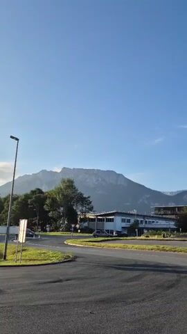 A wide shot captures a clear blue sky above a mountainous landscape. In the foreground, a paved road curves, with a traffic island on the left adorned with a lamp post and a white sign. A car is seen driving in the distance. Behind the road, a low-rise building complex stands, with trees interspersed. The scene appears to be an urban or suburban setting with a backdrop of prominent mountains.