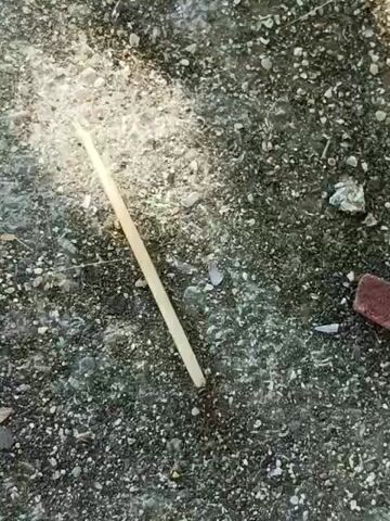 A close-up, overhead shot shows a wooden matchstick lying on a dark, gravelly surface. The matchstick is positioned diagonally across the frame, with its base embedded in the ground. A faint wisp of smoke is visible rising from the top of the matchstick, indicating it is either freshly lit or recently extinguished. The lighting suggests it is daytime.