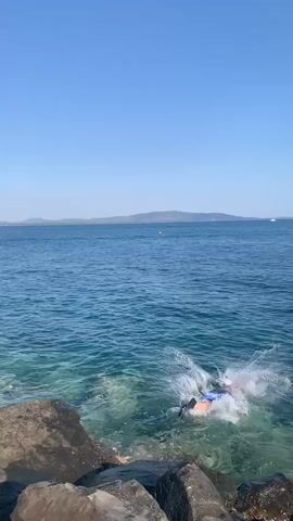 A person jumps into the ocean from rocks. The person is wearing a pink top and blue shorts. The water is clear and blue. The sky is clear and blue. There are hills in the distance.