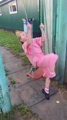 A young girl with blonde hair in braids, wearing a pink t-shirt and shorts, pink and white striped socks, and black crocs, is swinging on a swing set. She is upside down on the swing, with her legs in the air. In the background, there is a green building with a white window, a cardboard box, and some grass. The ground is covered with a gray mat. The scene appears to be outdoors, possibly in a backyard, with a wooden fence to the right. The girl seems to be enjoying herself, with a smile on her face. The weather appears to be daytime, possibly late afternoon, with natural lighting. The location is identified as Nizhnii Bugrysh, Russia. There is no visible text in the media file.