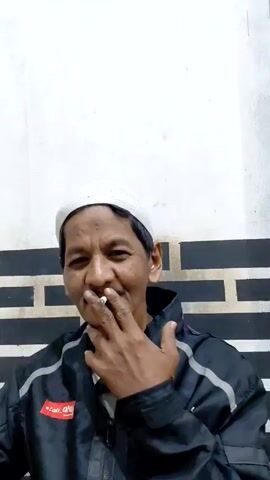 An adult man wearing a white skullcap, black jacket, and a mustache is holding a cigarette in his right hand and smoking it. Smoke emanates from the lit end of the cigarette. He has a slight smile on his face and is looking directly at the camera. The background is a plain, white wall with a horizontal black and white brick-like design toward the bottom. A red patch with white lettering is visible on the left side of his jacket.