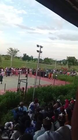 The video shows an outdoor event with a crowd of people gathered around a fenced-in court, possibly for a game or performance. The court is marked with red and white lines and has a net stretched across the middle. Several people are visible on the court, some appearing to be participating while others observe. The spectators are positioned both on elevated seating and on the ground surrounding the court. The environment appears to be a green, open area with trees in the background and a cloudy sky overhead. The time appears to be late afternoon or early evening, judging by the lighting conditions. Some people in the foreground are wearing traditional clothing, including hats.
