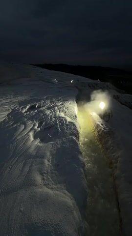 The video shows a narrow, shallow stream of water flowing through a white, rocky landscape at night. A bright light source illuminates the stream, creating a hazy effect as steam rises from the water. The surrounding terrain is uneven and textured, resembling a frozen or mineral-covered surface. The sky is dark, and the overall atmosphere is dimly lit.