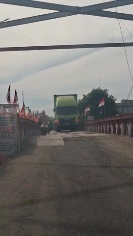 The video shows a view from behind a motorcycle driving across a bridge. There is a large green truck ahead of the motorcycle, also on the bridge. Several Indonesian flags are visible along the sides of the bridge, some attached to poles. A sign with text is also visible on the left side of the bridge. The sky is cloudy. The motorcycle the camera is behind is moving forward, as is the truck.