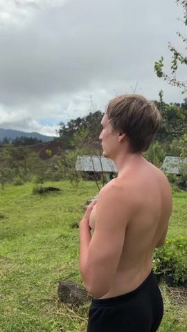 A shirtless man with blond hair stands in a grassy outdoor area, facing away from the camera and looking to the left. He is wearing black pants. His right hand is touching his chest. In the background, there are trees, hills, two small buildings with white roofs, and a cloudy sky. The man is in focus, while the background is slightly blurred.