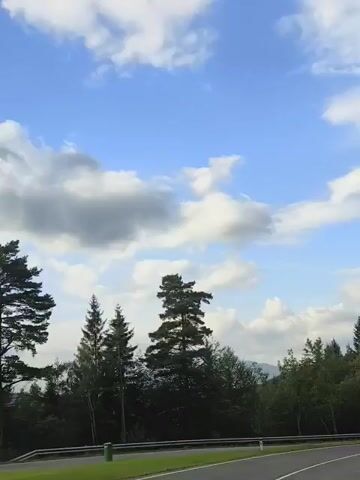 This is a vertical, low-angle video showing a winding road with a guardrail and trees. The sky is blue with scattered clouds. A sign is visible on the right side of the road.