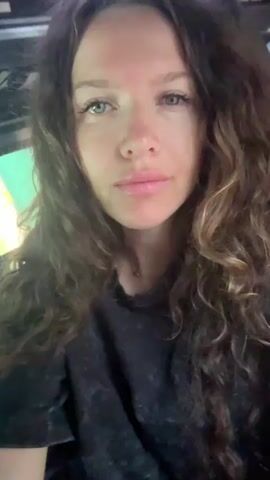 The video begins with a close-up of a woman with long, curly brown hair, her eyes closed and her lips slightly parted. The background is dark, with a hint of green light. The shot then quickly blurs and pans to the right, showing the woman's face in motion and a glimpse of trees outside a window. The scene changes to the interior of what appears to be a bus. The woman is now visible from the side, looking directly at the camera. Her eyes are open, and she has long, curly brown hair. Other passengers are visible in the blurred background, seated behind her. The woman is wearing a dark t-shirt. The environment suggests a journey on a bus, possibly during the day, given the glimpses of foliage through the windows.