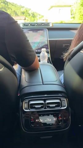 The video shows the interior of a car from a rear-seat perspective. A person's arm wearing a black shirt and blue jeans rests on the center console. A bottle of water and a smartphone are also visible on the console. The car's dashboard features a large touchscreen displaying a navigation map. The climate control panel for the rear passengers is visible below the air vents. In the front, a driver wearing sunglasses is reflected in the rearview mirror. The car is driving on a street lined with houses and trees under a clear blue sky. The scene appears to be a sunny day in a rural or suburban area.