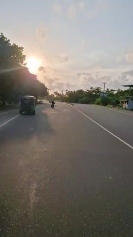 The video shows a road at dawn or dusk, with the sun low on the horizon. The road is paved and has painted white lines marking the lanes. There are trees and vegetation on either side of the road. Several vehicles are visible: a green tuk-tuk in the foreground, a car further down the road, and a motorcycle in the middle distance. The sky is bright, with some clouds. There are also power lines running alongside the road.