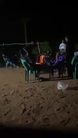 At night, on a sandy area with some vegetation, a group of people are gathered around tables and chairs. Some are seated, while others are standing or moving around. A man wearing a red hat and dark clothing is standing behind a person sitting in a green chair, appearing to interact with them closely. The scene is lit dimly, likely by artificial lighting.