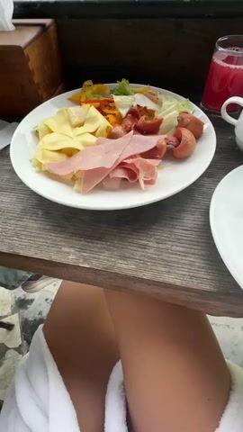 The video shows a breakfast spread on a wooden table with a breathtaking mountain view in the background. On the table, there's a plate containing what appears to be cheese slices, ham, and sausages, alongside a small salad. There's also a glass of red juice, a tissue box, and a plate of plums. The camera pans upwards to reveal a stunning vista of green mountains and valleys dotted with small villages, partially obscured by clouds. The perspective is from under a covered veranda, suggesting an outdoor dining setting at a mountain resort or lodge. A person's legs, clad in what looks like a white bathrobe, are visible under the table, indicating someone is seated there enjoying the meal and the scenery.