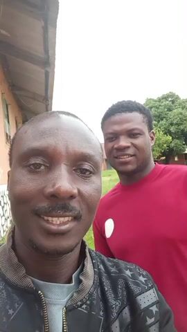 This is a selfie-style video featuring two Black men standing outside. The man in the foreground, on the left, is closer to the camera and smiling. He has a mustache and is wearing a black jacket with a grey shirt underneath. The man in the background, on the right, is also smiling and wearing a red shirt with a white oval logo on it. They appear to be in front of a building with windows and a corrugated roof. Trees and a grassy area are visible in the background. The lighting suggests it is daytime.